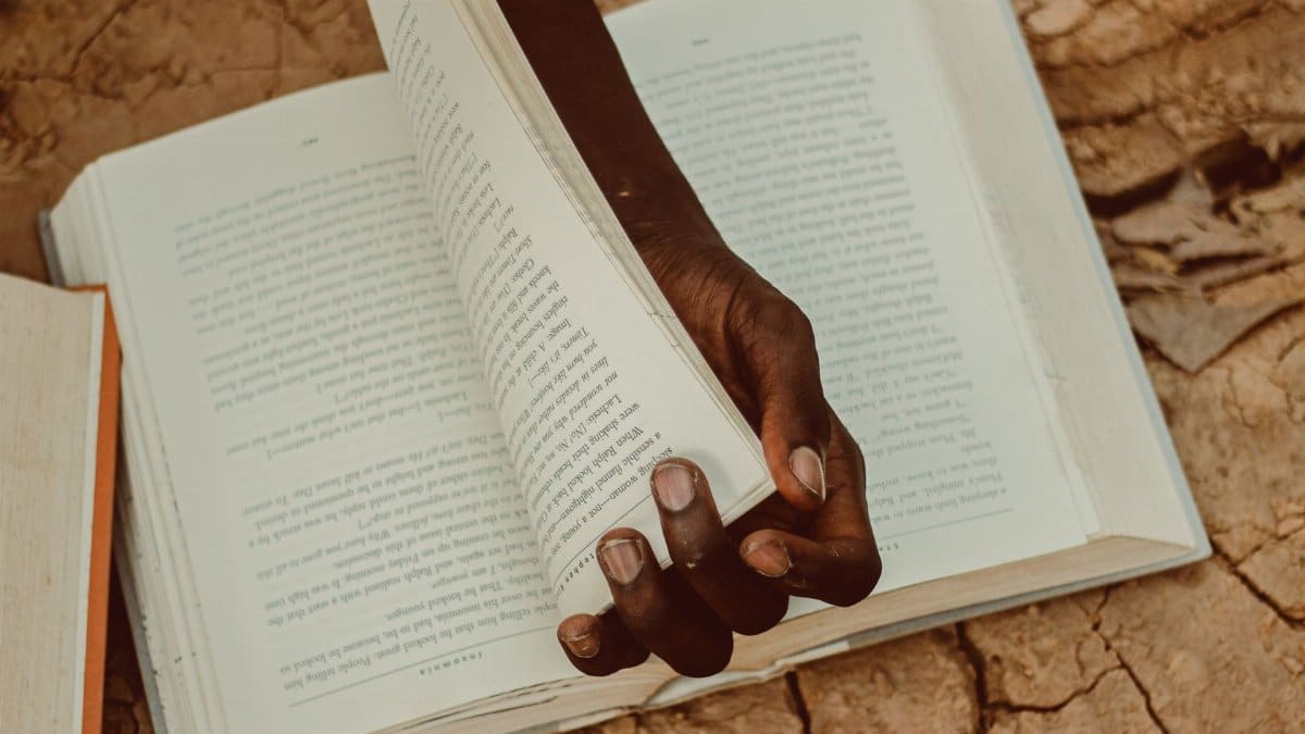 A person holds a book open on parched, cracked earth, symbolizing knowledge amidst adversity.