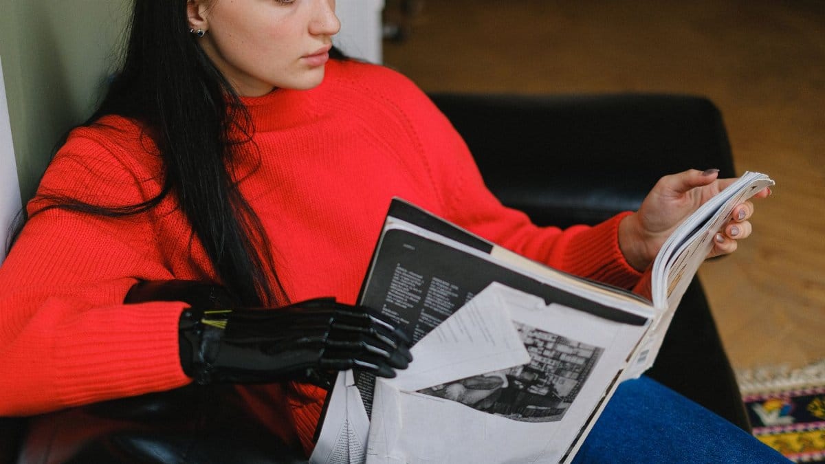 Woman with a prosthetic hand in a red sweater reading a magazine indoors.