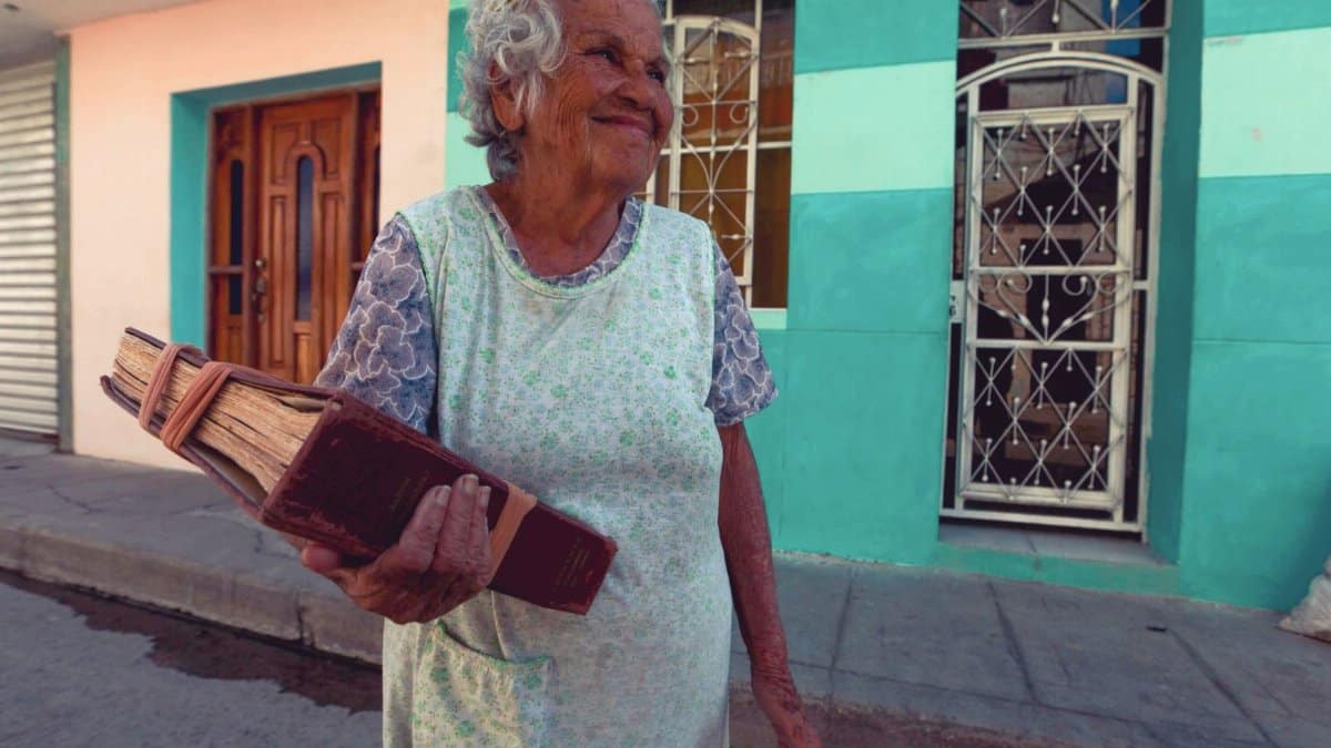 Joyful elderly woman holding a large book while walking down a colorful street.