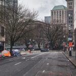 A city street with tents showing urban homelessness beneath bare trees on cloudy day.
