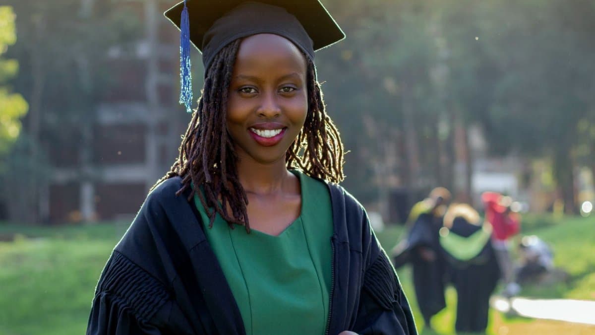 A young woman wearing a graduation cap and gown, smiling outdoors on campus.
