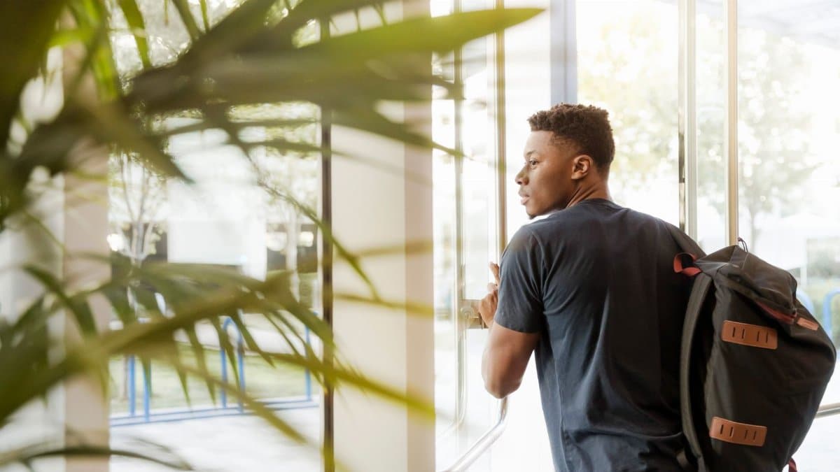 A college student with a backpack exits a campus building through glass doors.
