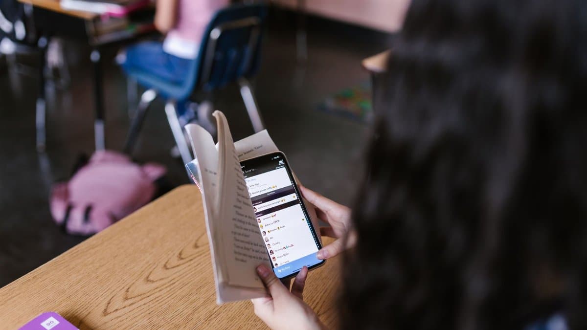 A student in a classroom multitasks by reading a book while using a smartphone, emphasizing modern educational challenges.
