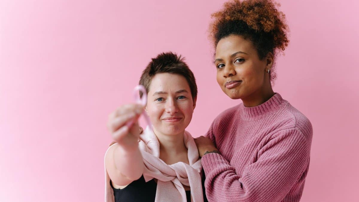 Two women embracing, one holding a pink ribbon for breast cancer awareness, against a pink background.