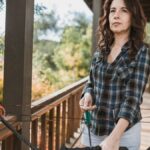 Woman holding a compound bow on a porch, ready to shoot outdoors.