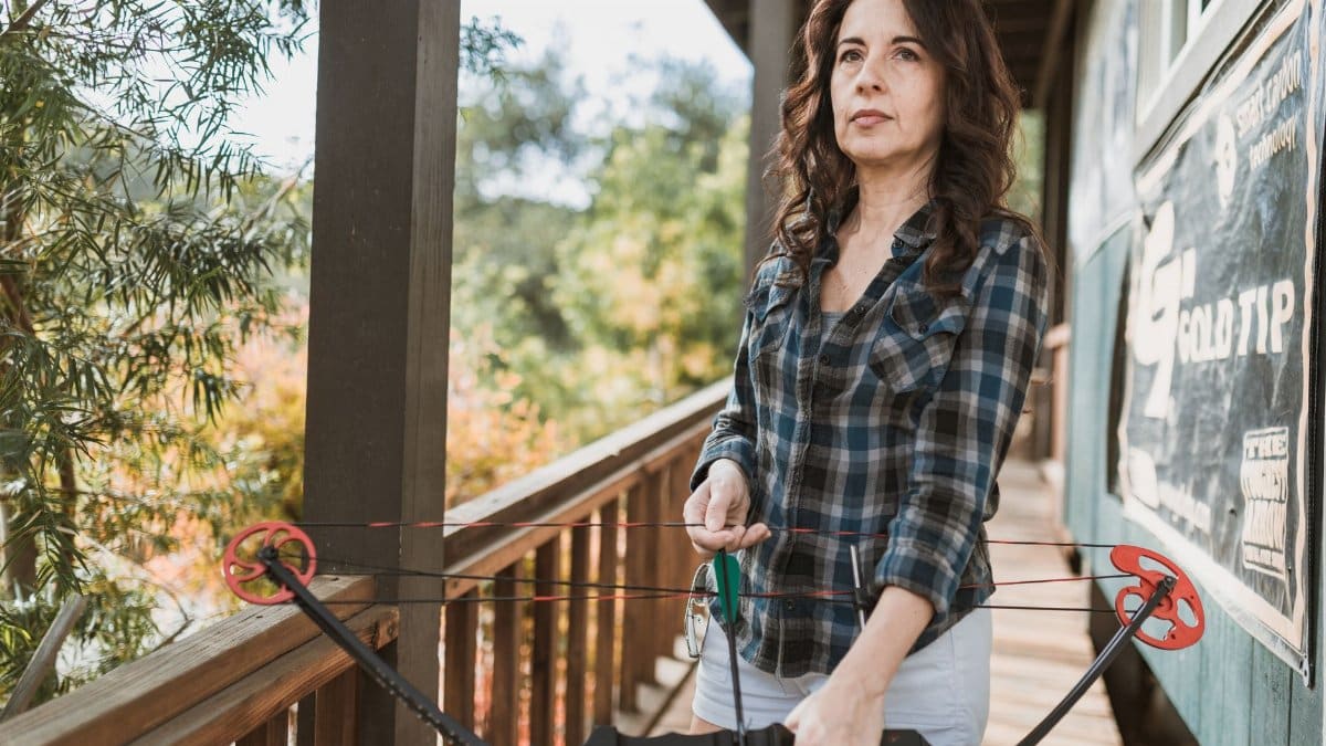 Woman holding a compound bow on a porch, ready to shoot outdoors.