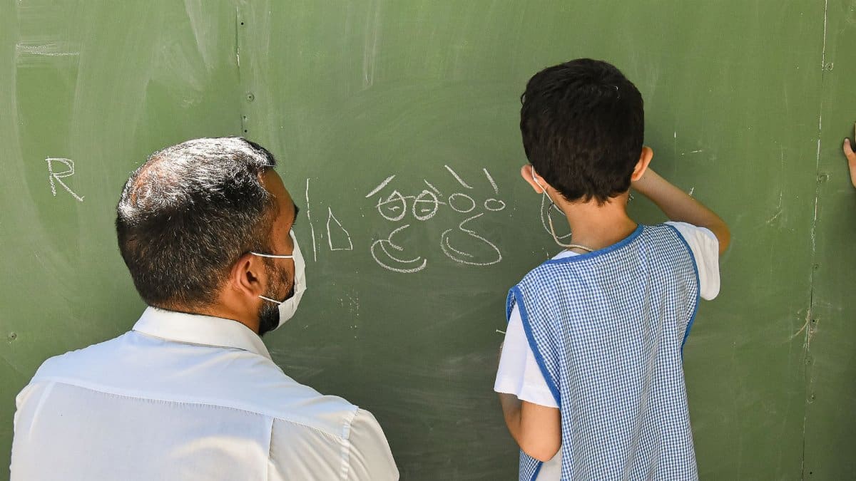 A teacher guides a student writing on a green chalkboard in a classroom setting.