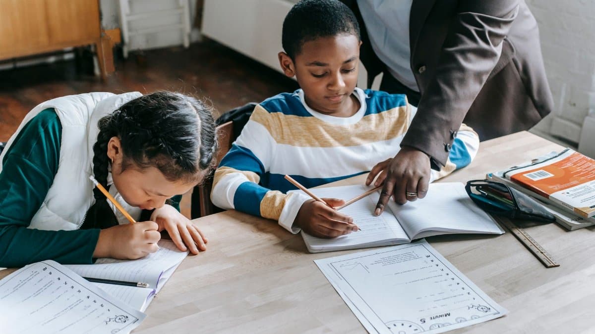 High angle of crop unrecognizable black female teacher explaining task to focused little schoolboy sitting at desk near attentive Asian classmate writing in notebook