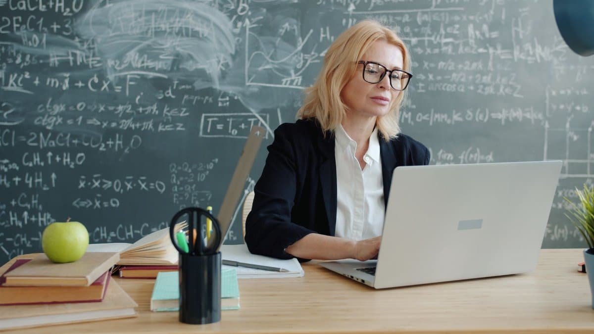 Teacher working on a laptop in a classroom with chalkboard equations, focused and professional.