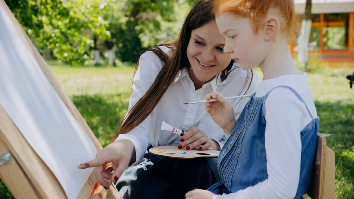 A teacher guides a child in an outdoor painting session, fostering creativity.