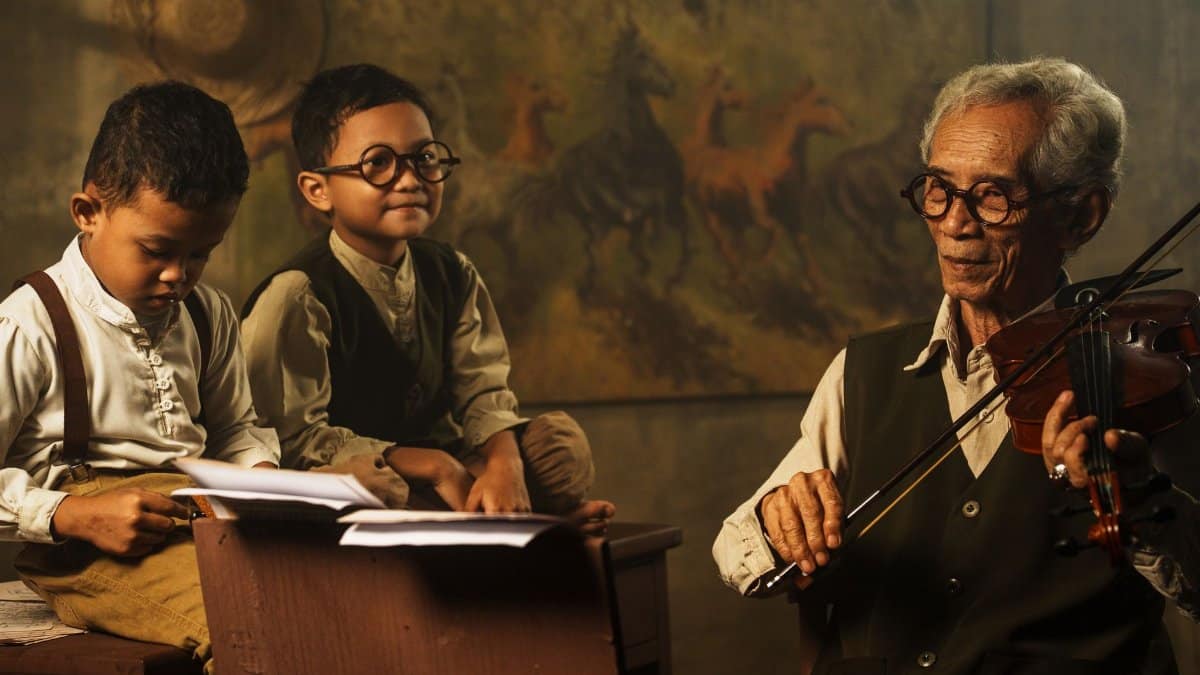 An elderly man plays violin while teaching two children indoors with warm lighting.