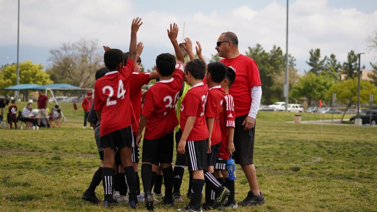A youth soccer team in red jerseys gathers in a huddle with their coach on an outdoor field.