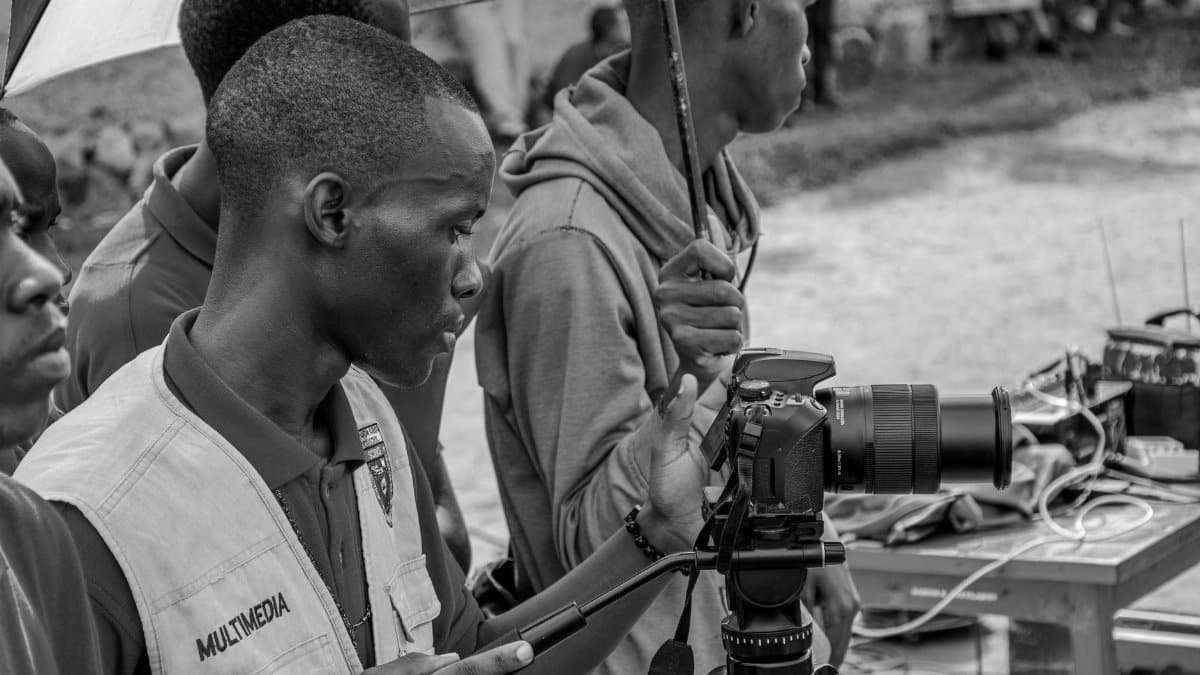 Black and white image of a filmmaker setting up a camera outdoors, capturing a documentary scene.