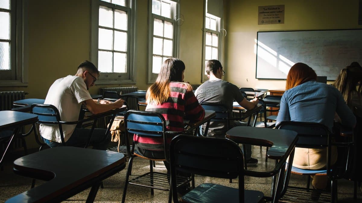 A group of students taking an exam in a classroom, illuminated by natural sunlight from windows.