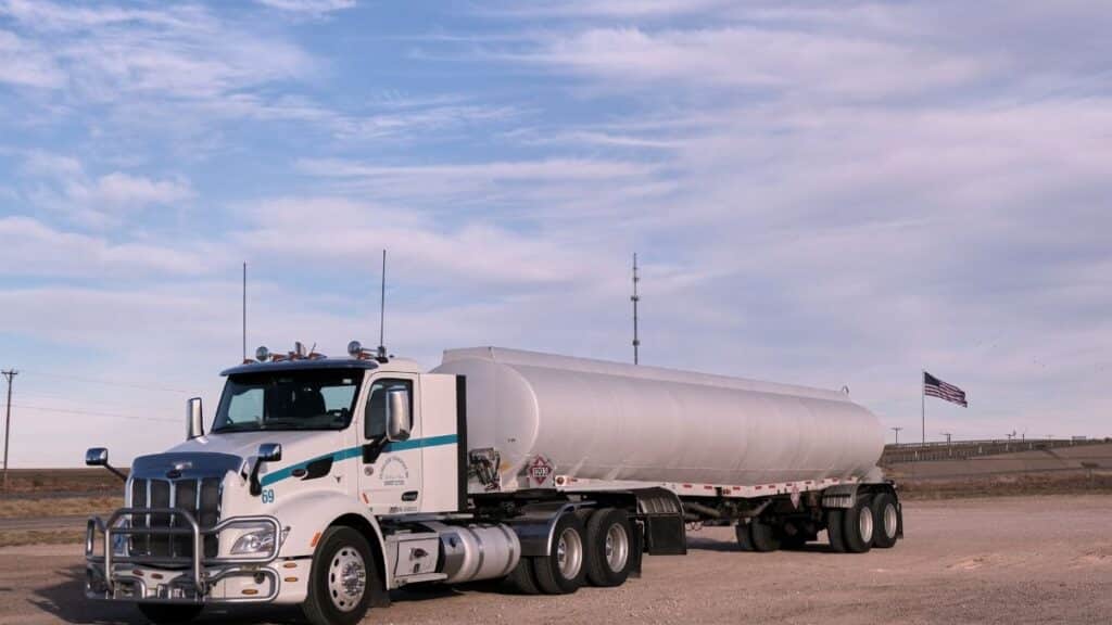 White fuel transport truck parked in a Texas landscape under clear skies, showcasing American flag in the distance.