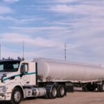 White fuel transport truck parked in a Texas landscape under clear skies, showcasing American flag in the distance.