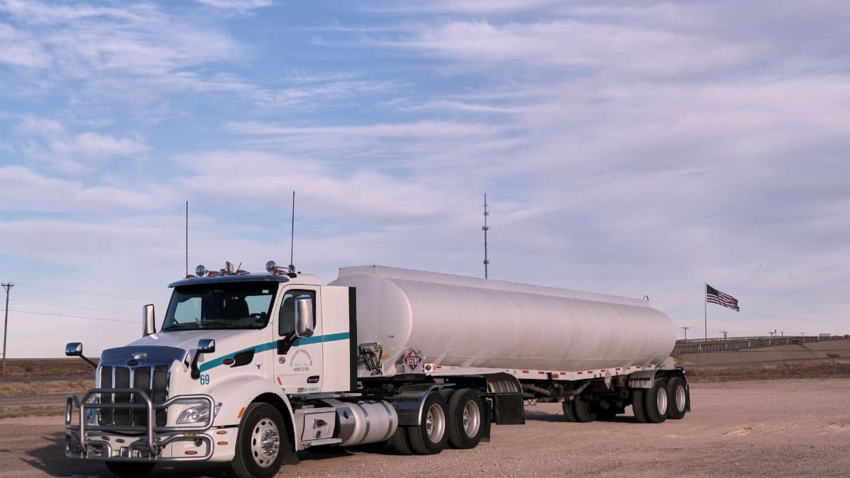 White fuel transport truck parked in a Texas landscape under clear skies, showcasing American flag in the distance.