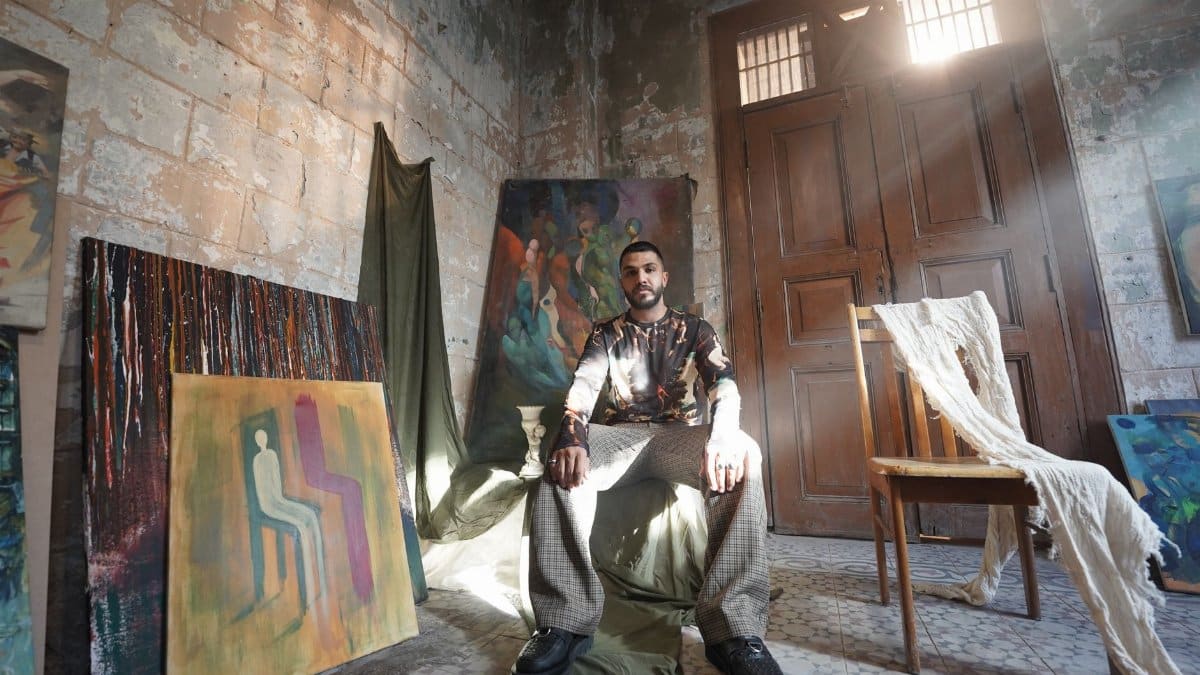 An artist sits among his paintings in a rustic, sunlit workshop, surrounded by art.
