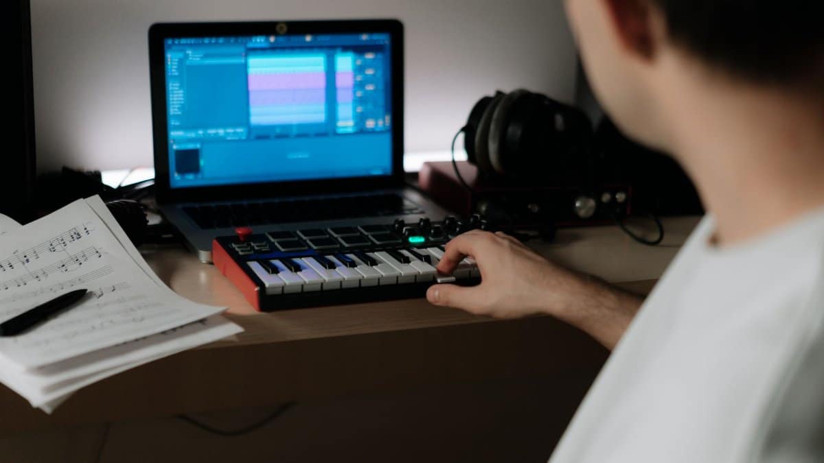 A music producer uses a synthesizer and laptop for composing in a home studio.