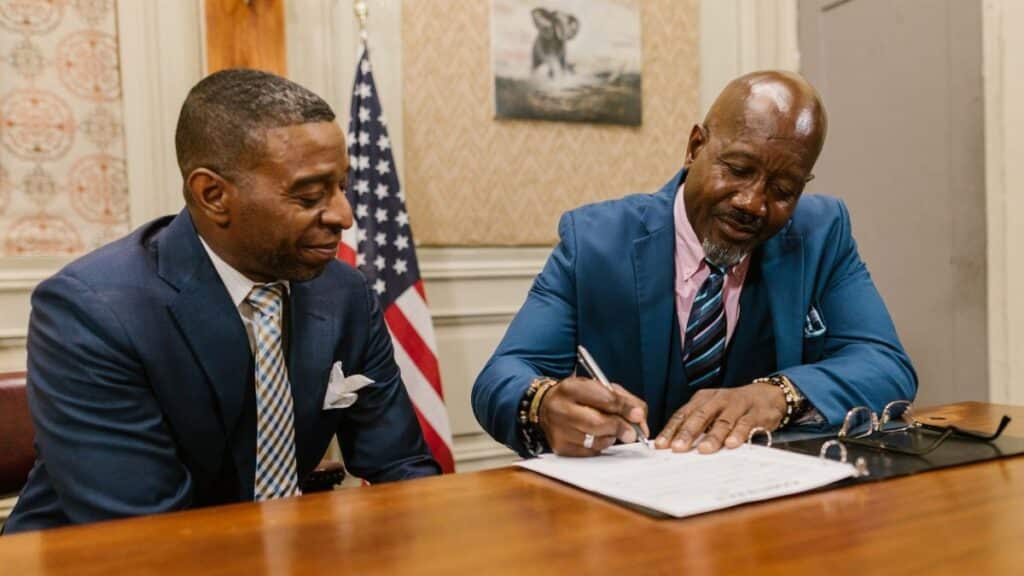 Two businessmen in suits signing documents in an office setting with an American flag.