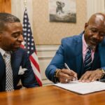 Two businessmen in suits signing documents in an office setting with an American flag.