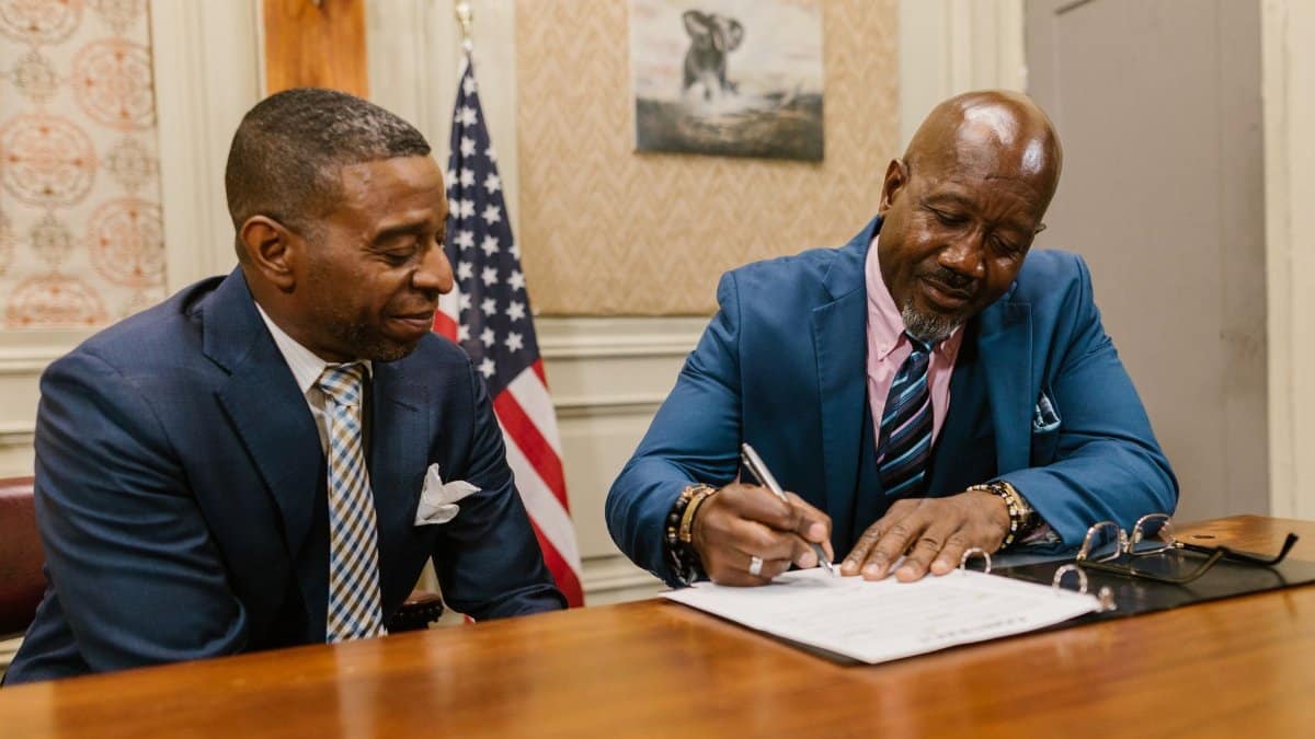Two businessmen in suits signing documents in an office setting with an American flag.