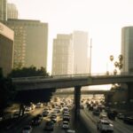 Heavy traffic on a Los Angeles freeway at sunset with buildings and palm trees in view.