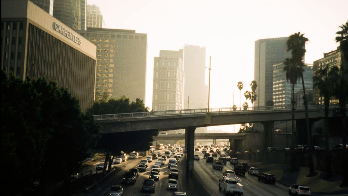 Heavy traffic on a Los Angeles freeway at sunset with buildings and palm trees in view.