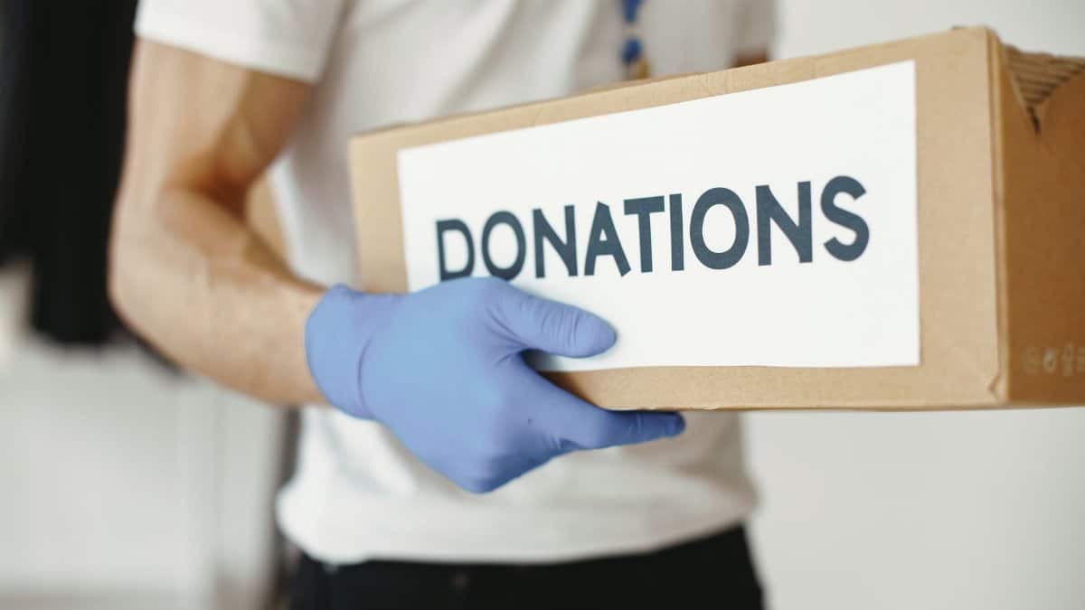 A volunteer wearing gloves holds a 'donations' box, symbolizing community support.