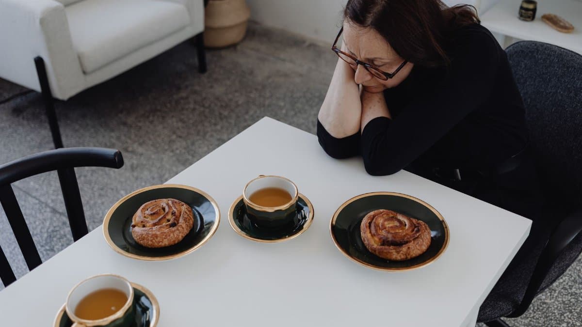 Thoughtful moment of a woman with eyeglasses indoors at a table with coffee and pastries, capturing solitude and contemplation.