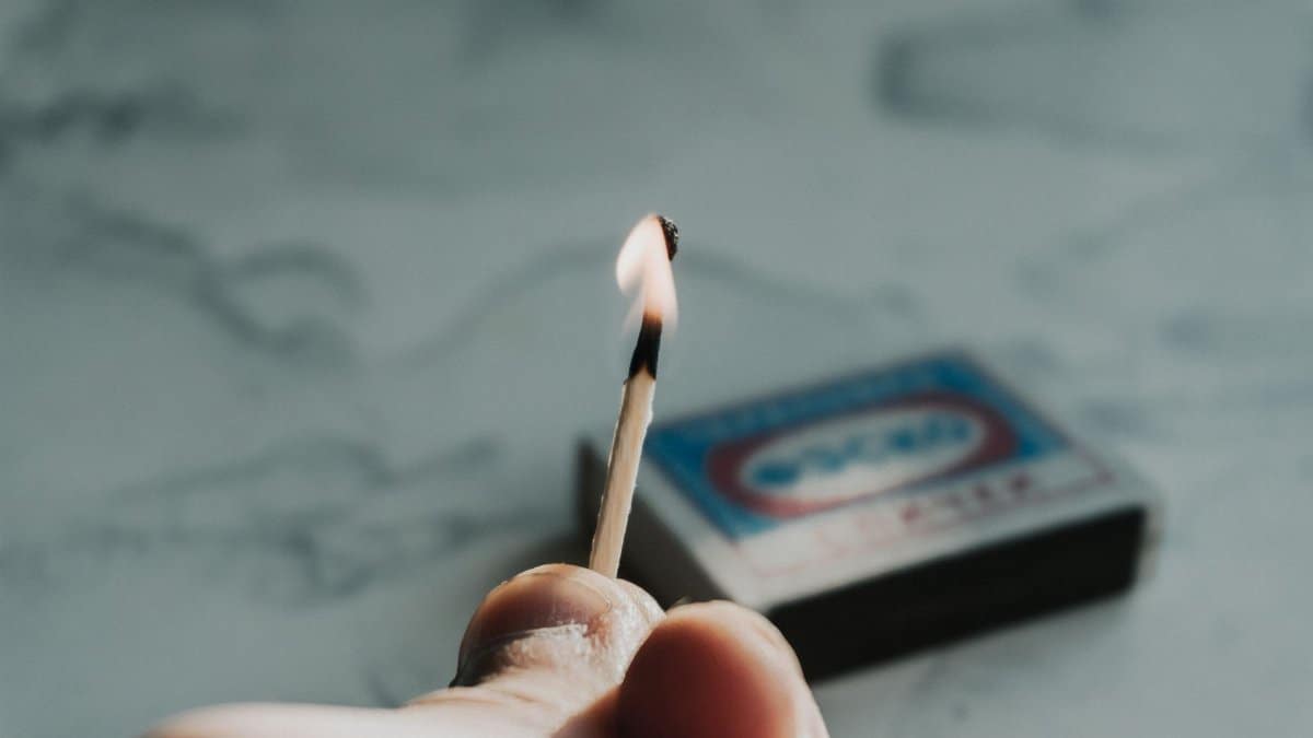 A detailed close-up of a hand holding a burning matchstick against a soft focus background.