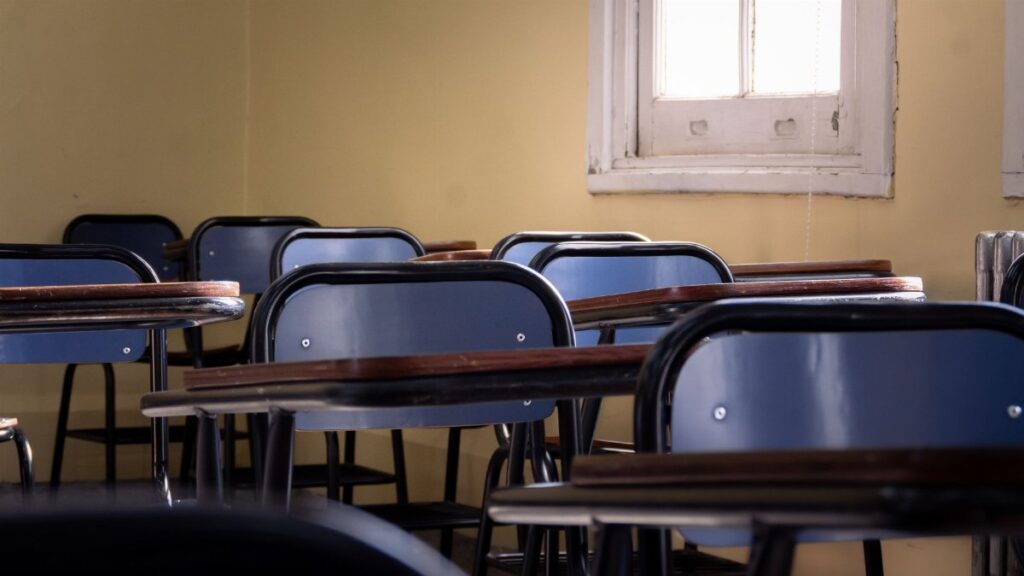 Row of empty blue chairs and wooden desks in a sunlit classroom with yellow walls.