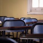 Row of empty blue chairs and wooden desks in a sunlit classroom with yellow walls.