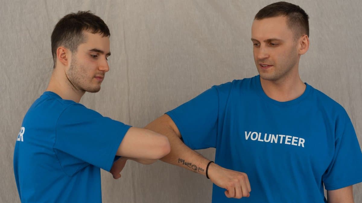Two male volunteers in blue shirts exchange an elbow bump, symbolizing support and teamwork.