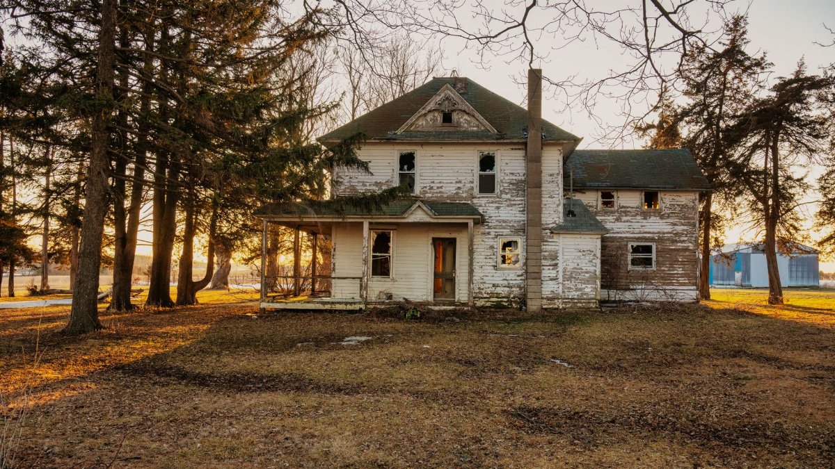 An old, weathered farmhouse stands amidst tall trees, lit by a warm sunset in Minnesota, USA.