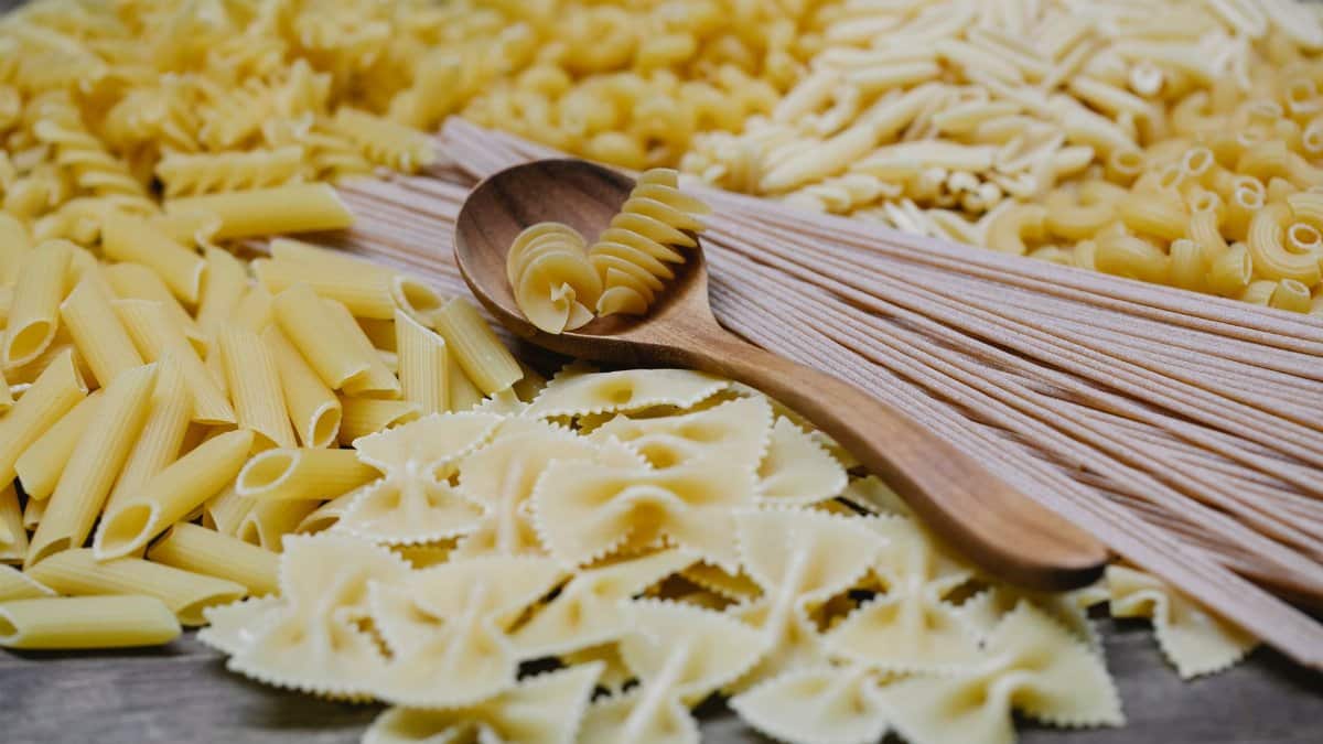 Arrangement of uncooked various pasta including spaghetti fusilli farfalle and penne heaped on table with wooden spoon
