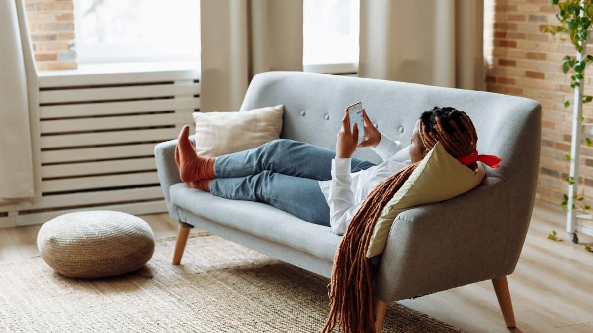 African American woman with dreadlocks relaxing and using phone on a modern sofa.