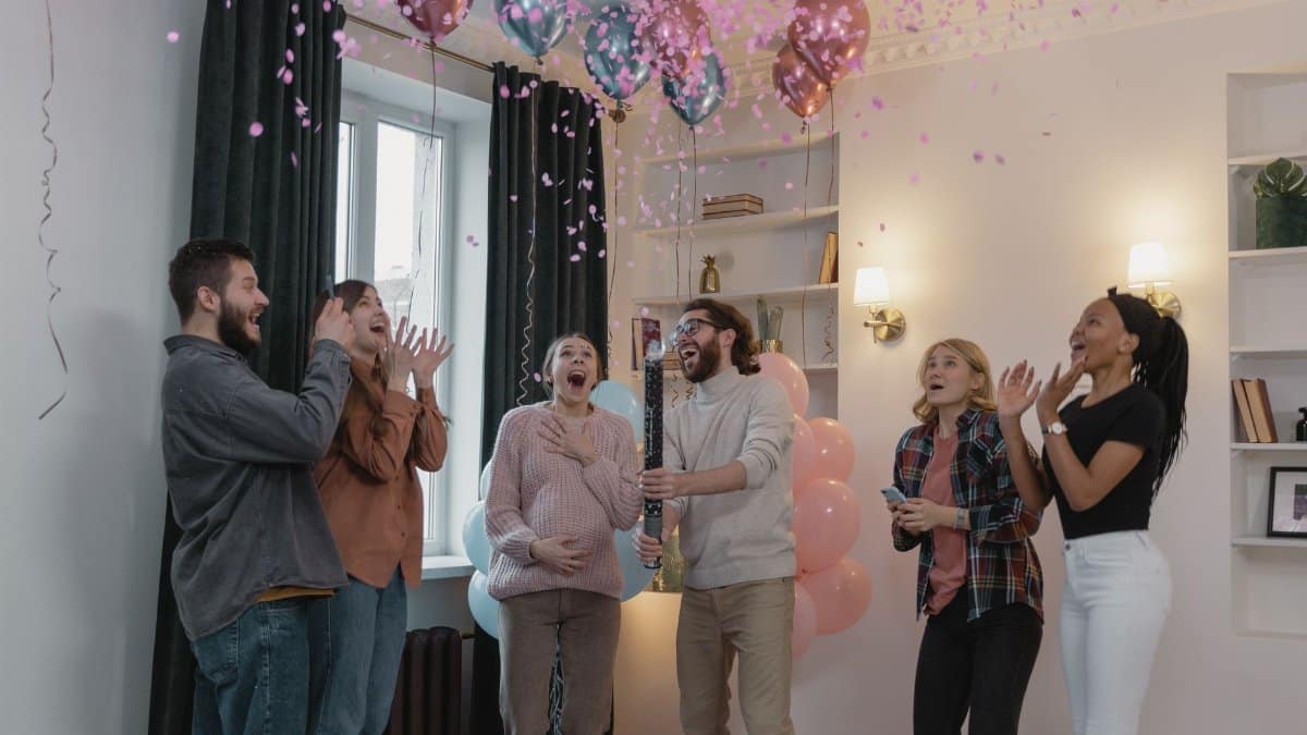 A happy group of friends celebrating a gender reveal party with pink confetti and balloons indoors.