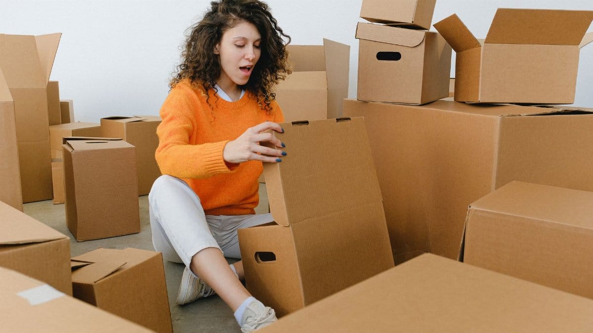 Amazed young female with curly hair in casual clothes sitting on floor and unpacking carton boxes with opened mouth after relocation