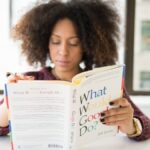 A woman reading 'What Would Google Do?' at a desk by a window in a modern office.