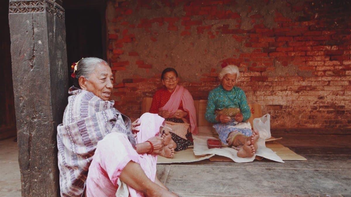 Three elderly women sitting on a porch with traditional clothing, engaged in a relaxed conversation.