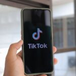 A hand holds a smartphone displaying the TikTok app in an indoor setting.