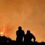Two people silhouetted in front of a large forest fire with intense flames and smoke.