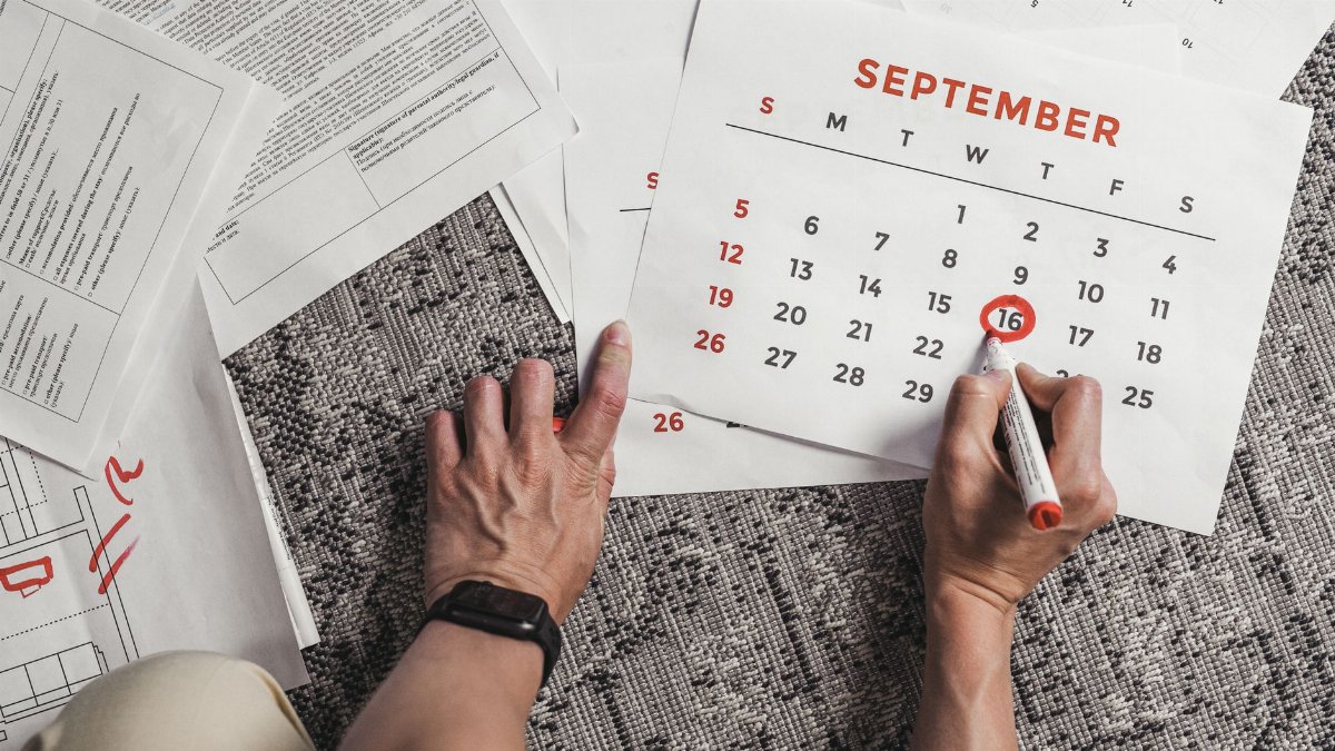 Hands holding a calendar with September dates encircled, surrounded by documents on a carpet.