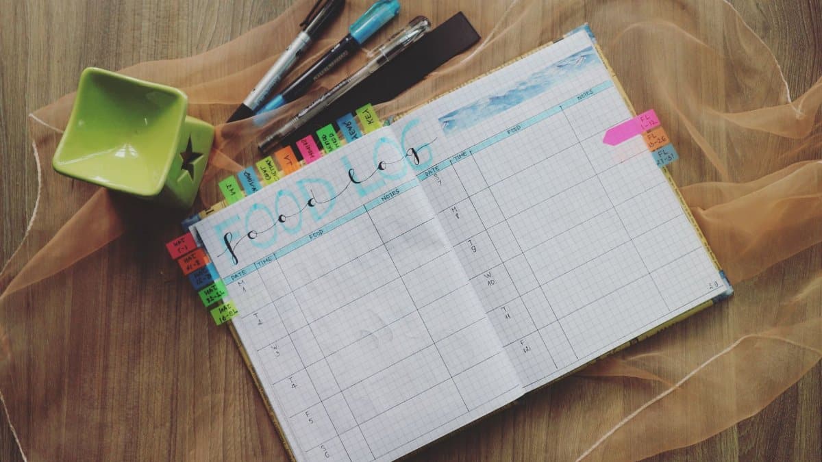Open food log planner with colorful tabs and pens on a wooden desk, perfect for meal tracking.