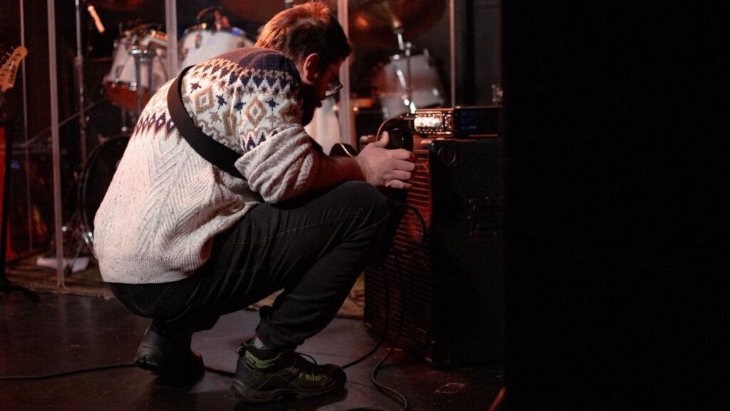 A musician wearing a patterned sweater tunes an amplifier on stage before a live performance.