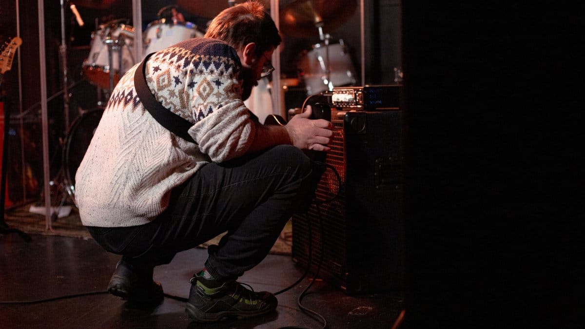A musician wearing a patterned sweater tunes an amplifier on stage before a live performance.