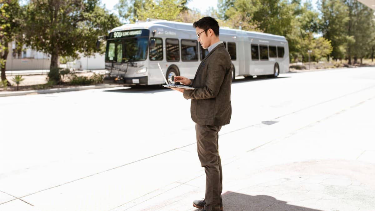 Businessman in corporate attire using laptop outdoors near bus stop, signifying remote work flexibility.