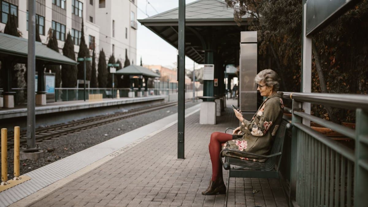Senior adult woman using a smartphone while sitting on a bench at an outdoor train station.