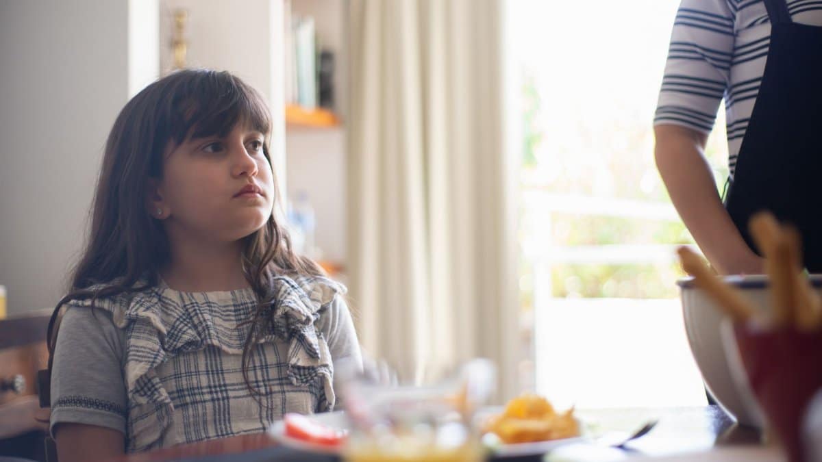 A young girl in a plaid shirt sitting indoors at a table, appearing thoughtful.